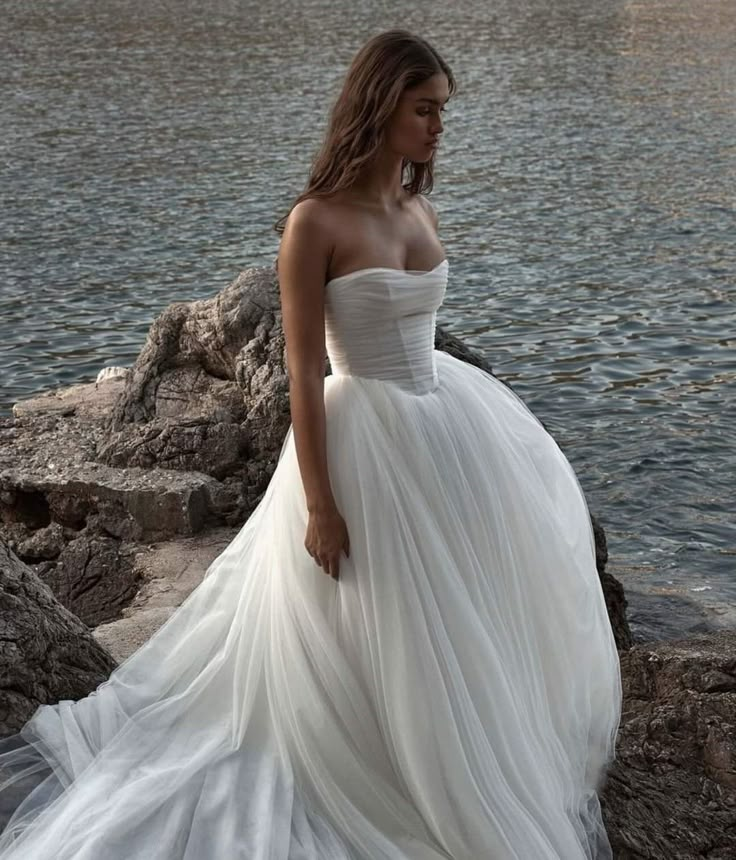 a woman in a white dress standing on rocks by the water with her hands behind her back