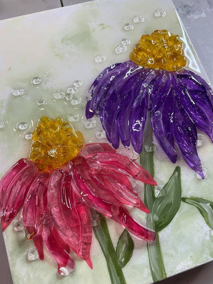 two colorful flowers sitting on top of a white table next to water droplets and green leaves