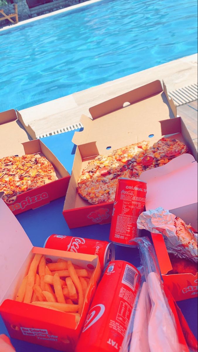 pizzas and french fries are sitting on the table by the swimming pool, ready to be eaten