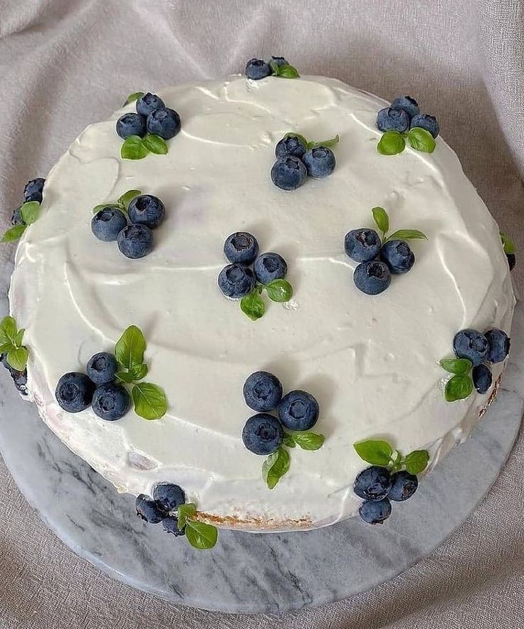 a white frosted cake topped with blueberries and green leaves on top of a table