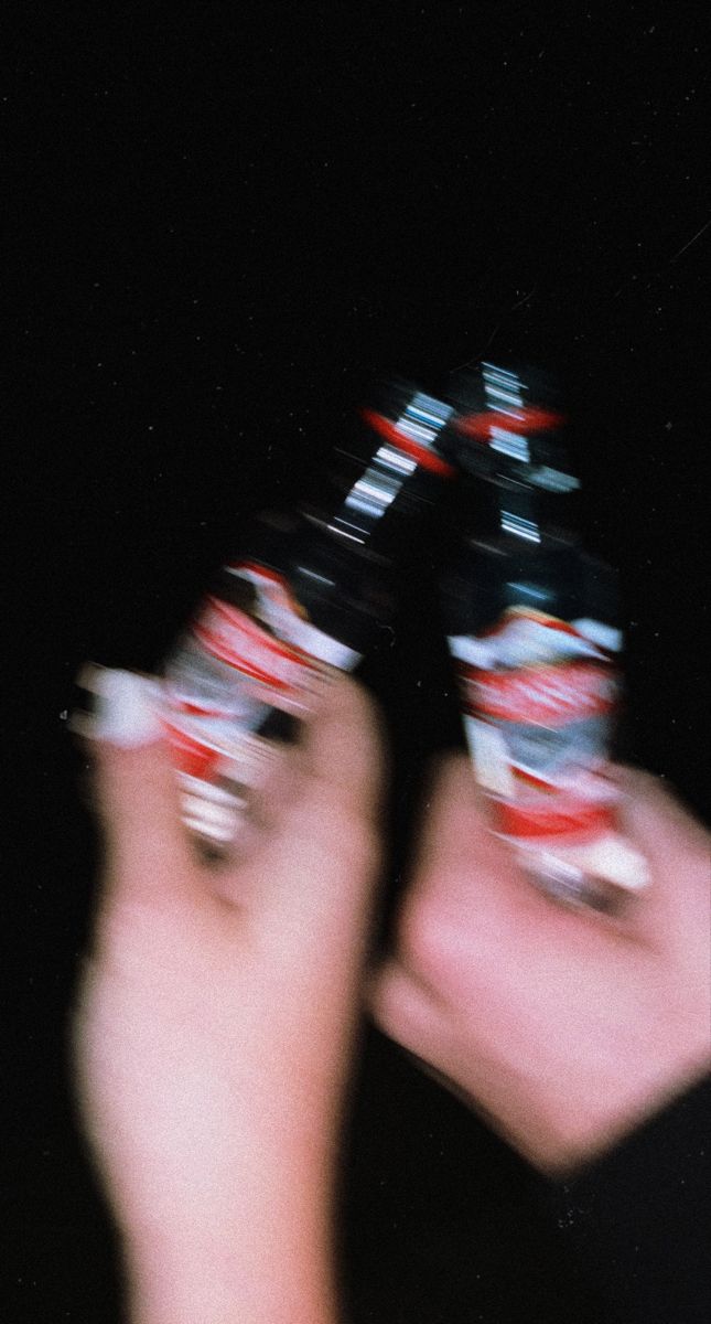 two hands holding up several soda cans in the dark, with blurry image behind them