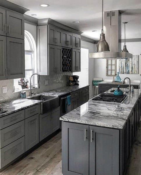 a large kitchen with gray cabinets and marble counter tops is pictured in this image, there are two pendant lights hanging from the ceiling