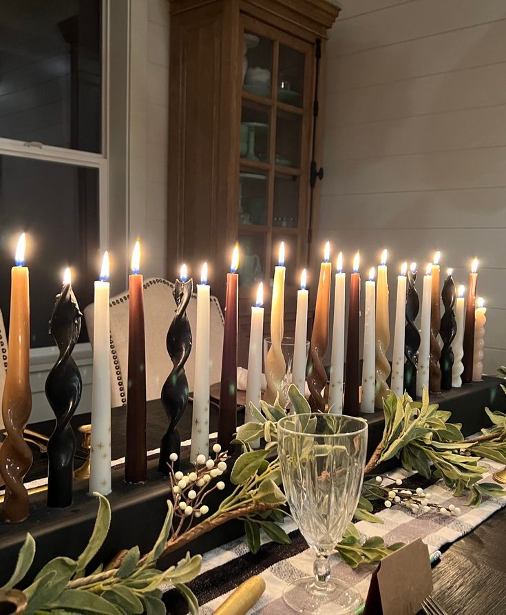 many candles are lit on a table with greenery and other decorations around it in front of a window