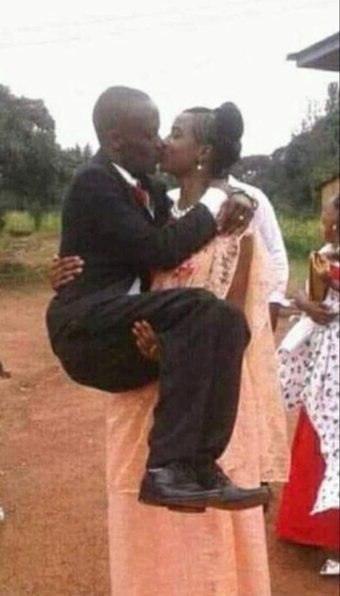 a man and woman kissing in front of people on a dirt road with trees behind them