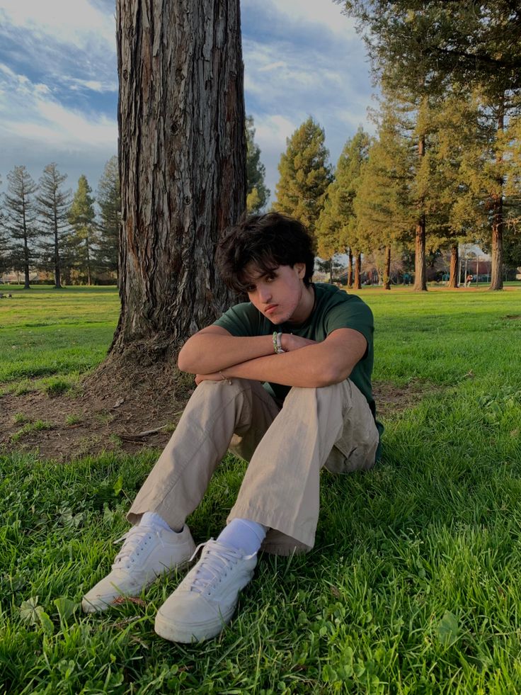 a young man sitting in the grass next to a tree with his arms crossed and head resting on his knees