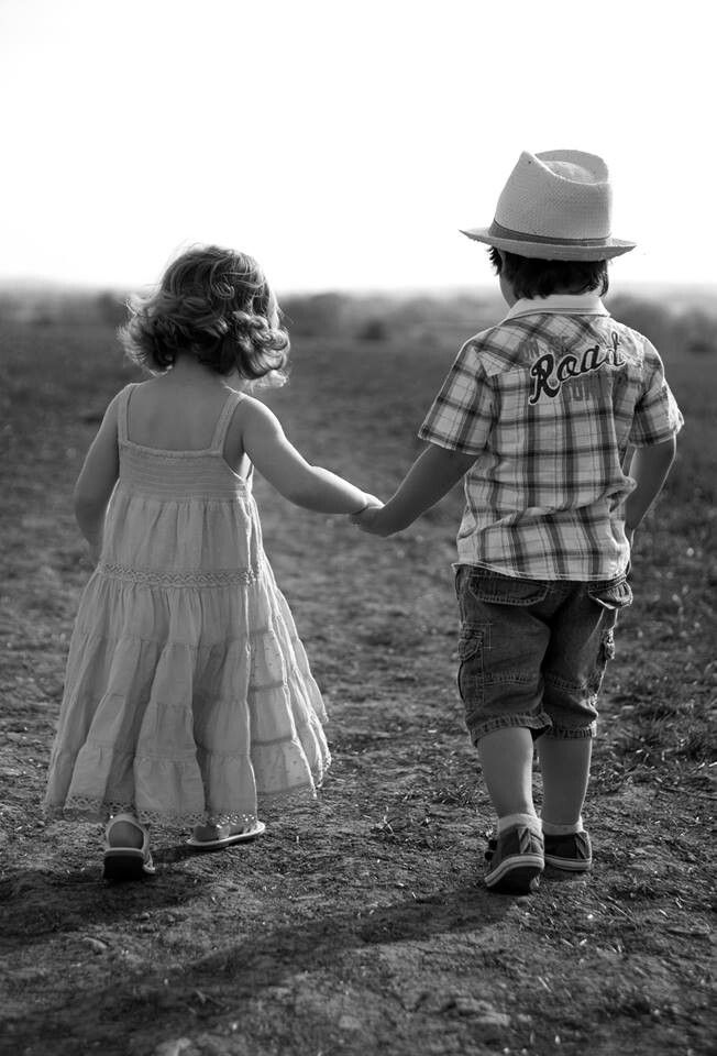 two young children holding hands in the middle of an open field, one is wearing a hat and the other wears a dress