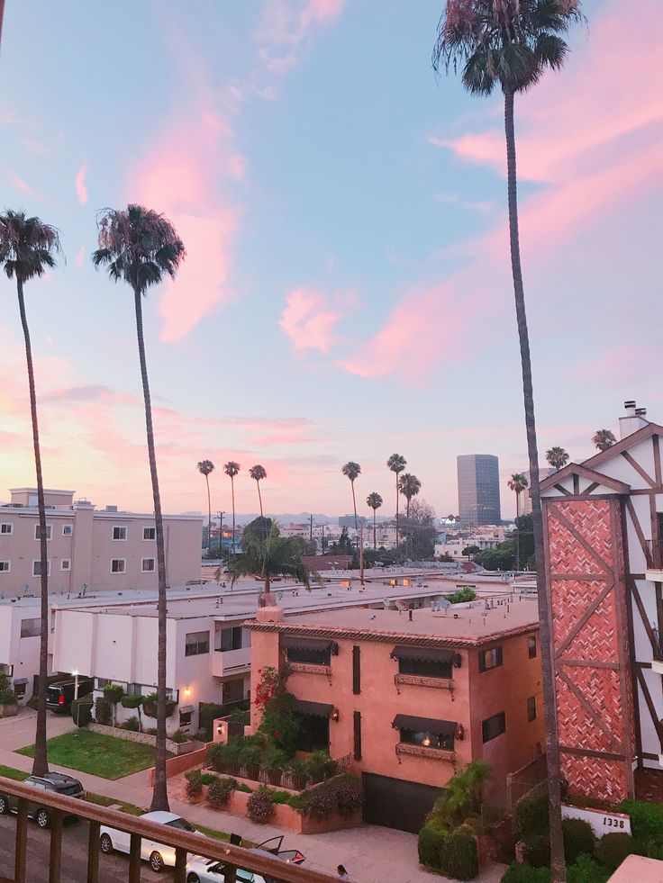 palm trees line the street in front of an apartment building at sunset with pink clouds