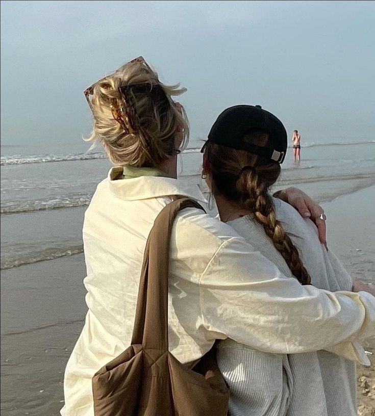 two people standing on the beach with their arms around each other looking out at the ocean