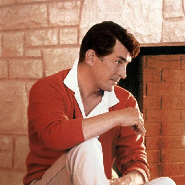 a man sitting on the floor in front of a fire place wearing a red sweater and white pants