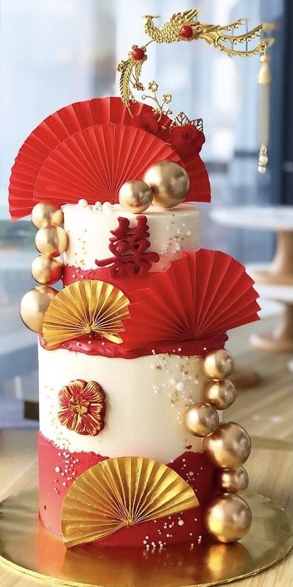 a three tiered cake with red and gold decorations on the top, sitting on a table