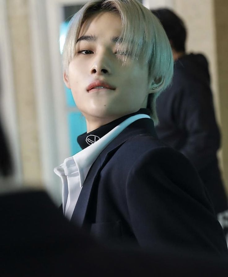 a young man with blonde hair wearing a black suit and white shirt, looking off into the distance