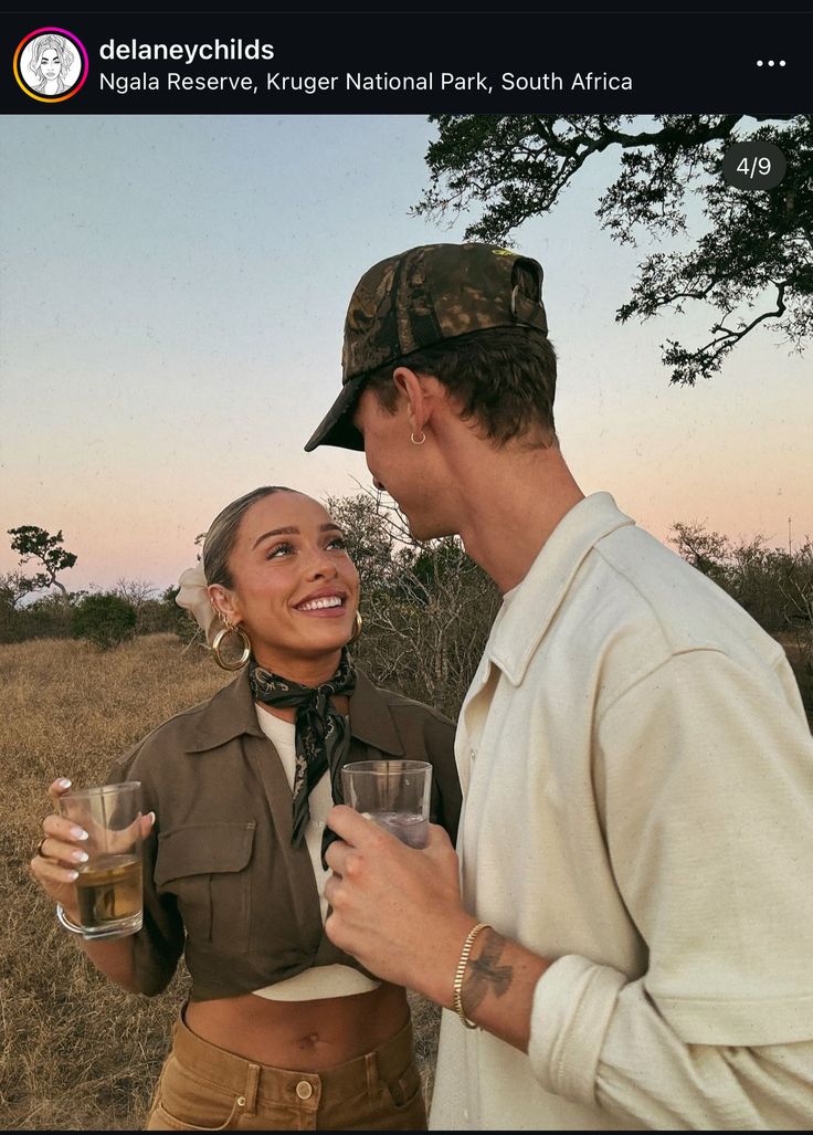 a man and woman standing next to each other holding wine glasses in their hands while the sun is setting