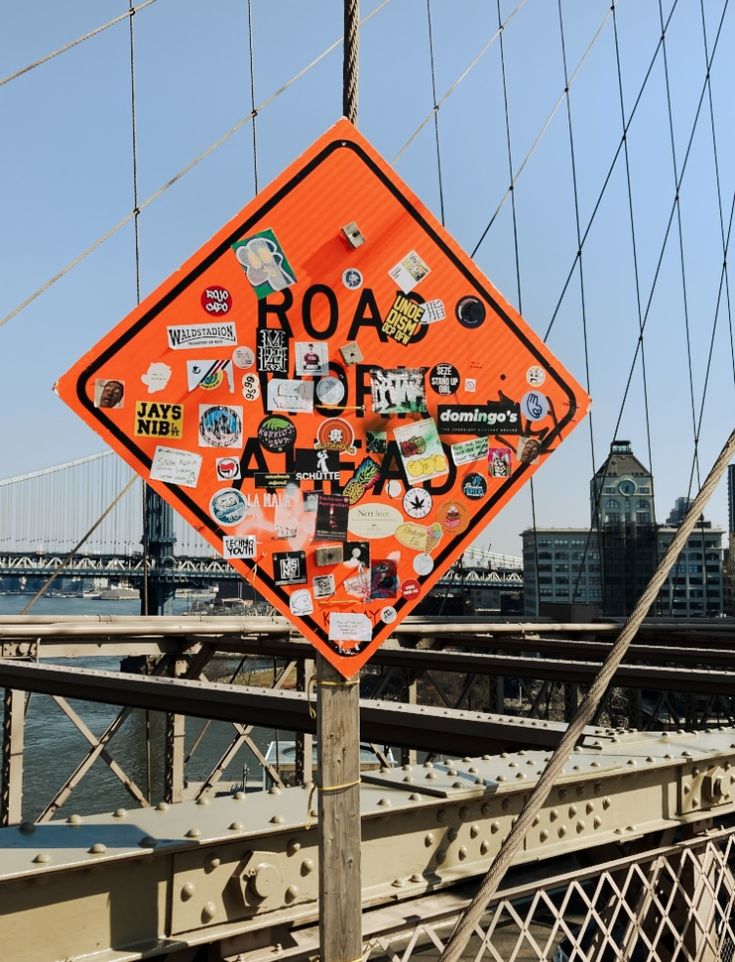 an orange road sign covered in stickers on the side of a bridge over water