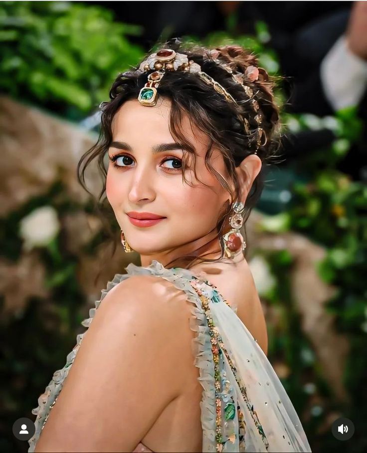 a woman with braids and jewelry on her head looking at the camera while standing in front of greenery