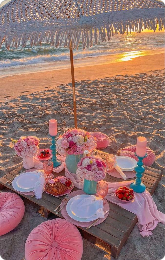 a table set up on the beach with plates and flowers in front of an umbrella