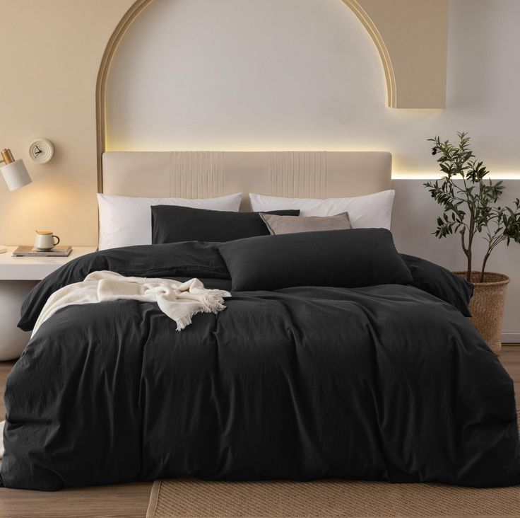 a bed with black linens and pillows in a white room next to a potted plant