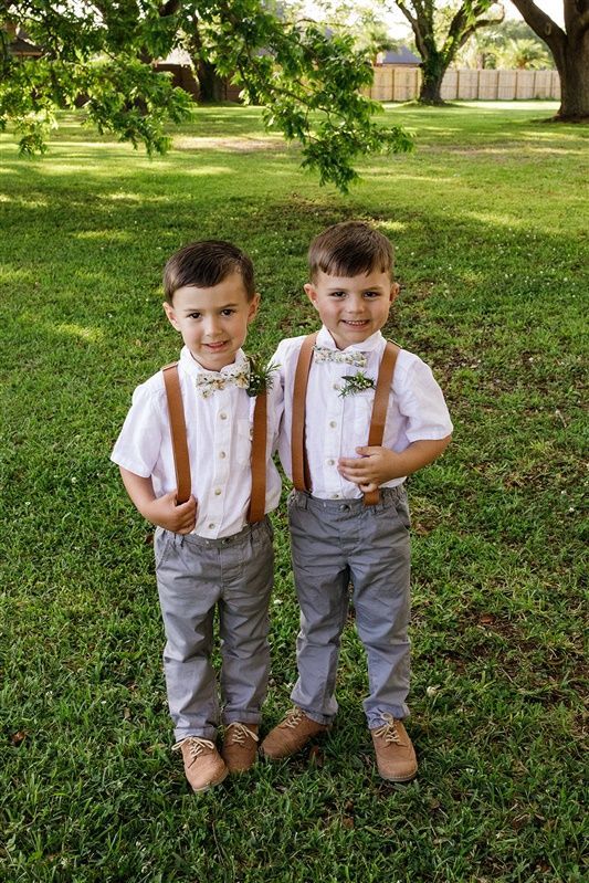 two young boys wearing suspenders and bow ties standing in the grass with trees behind them