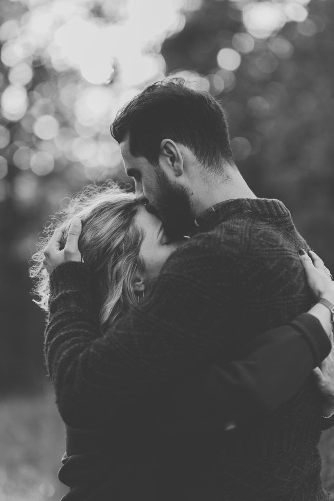 black and white photo of a couple embracing each other in front of some trees with the sun shining on them