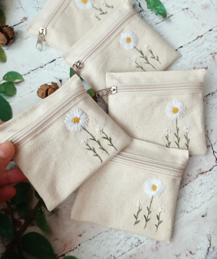 three small zippered pouches with daisies embroidered on the side and one being held by a person's hand