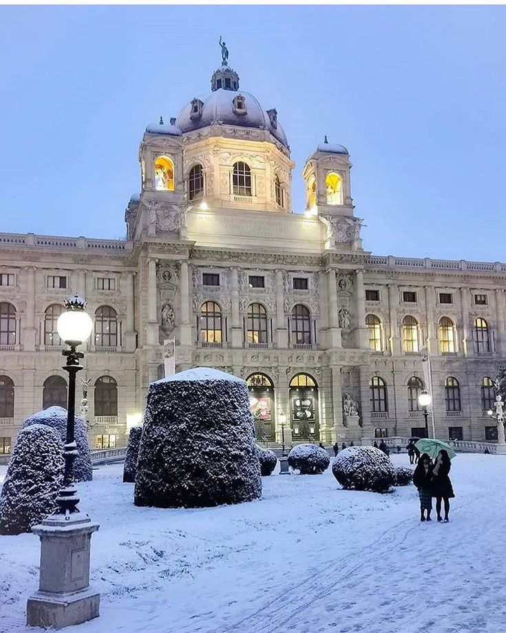 two people walking in front of a large building with snow on the ground and trees