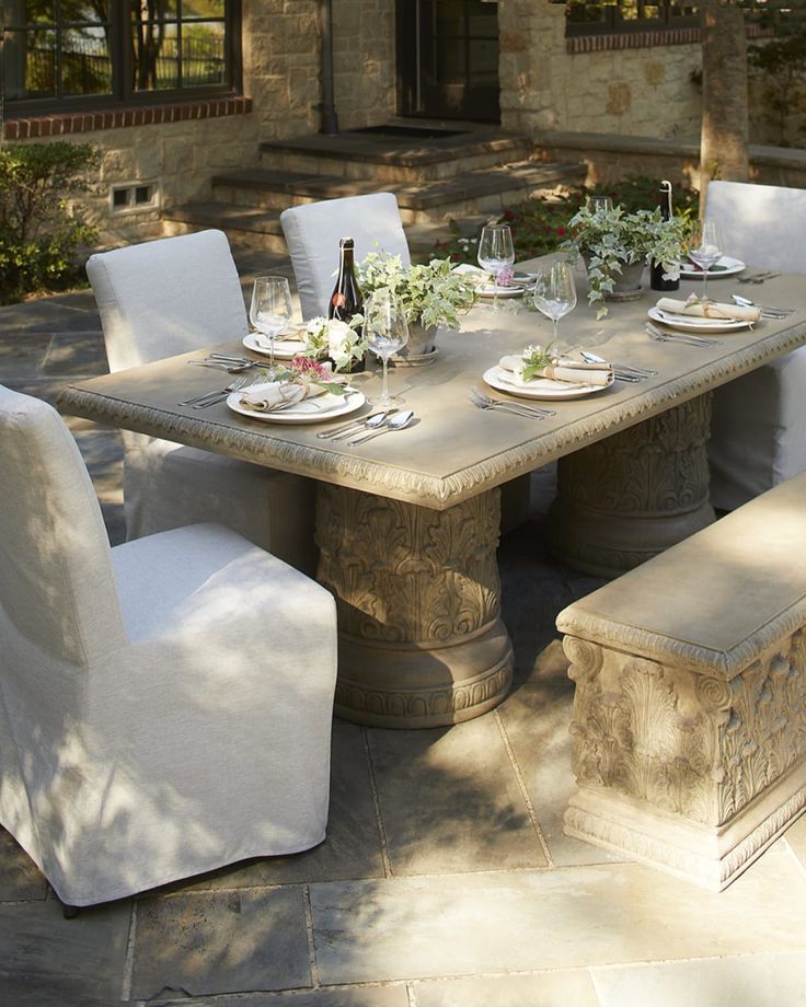 an outdoor dining table with white chairs and place settings on it, in front of a stone building