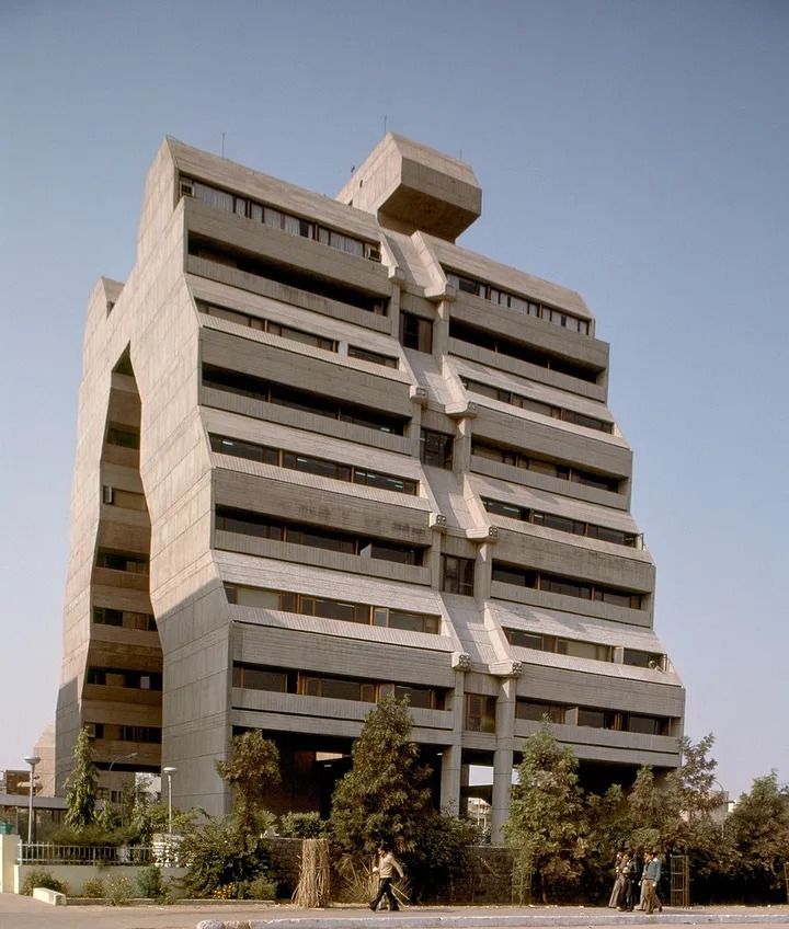 an unusual building in the middle of a parking lot with people walking around and on bicycles