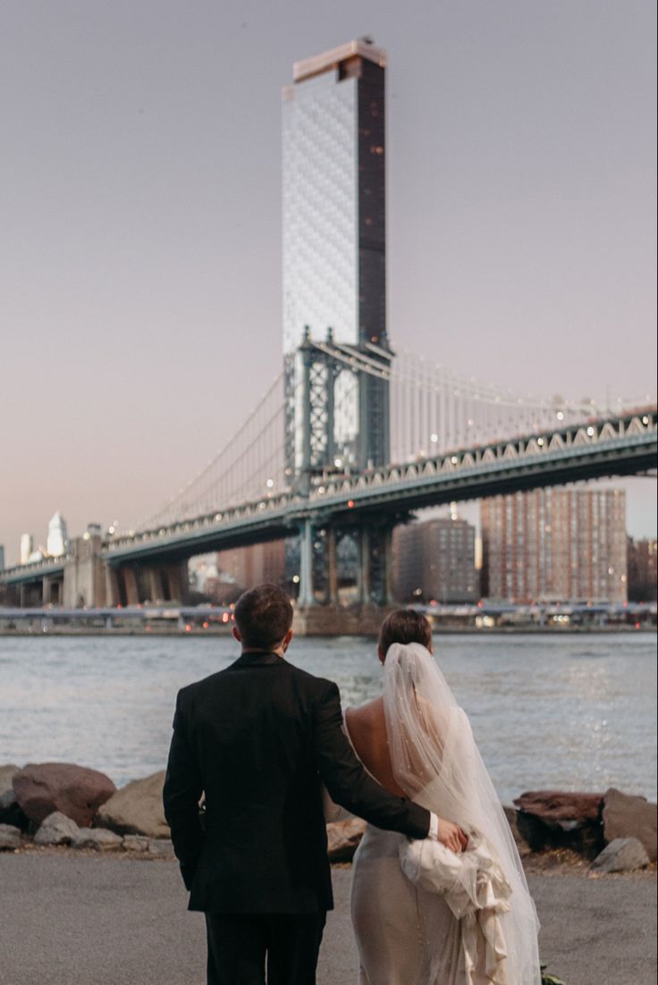 the bride and groom are walking by the water in front of the brooklyn bridge on their wedding day