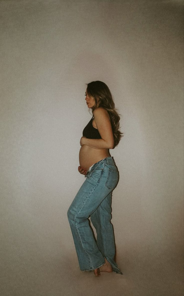 a pregnant woman in jeans is posing for the camera while holding her belly up and looking down