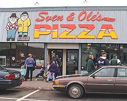 cars parked in front of a pizza shop with people walking around the building and on the sidewalk