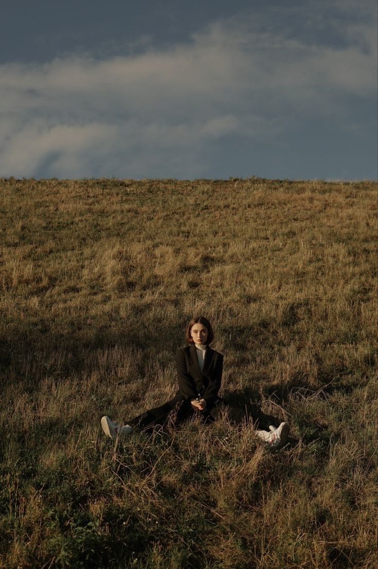 a woman sitting on top of a grass covered field next to a bird in the sky