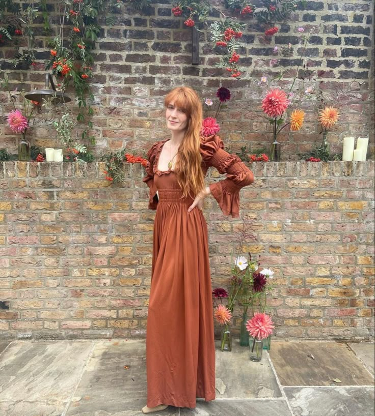 a woman standing in front of a brick wall wearing an orange dress and holding her hands on her hips