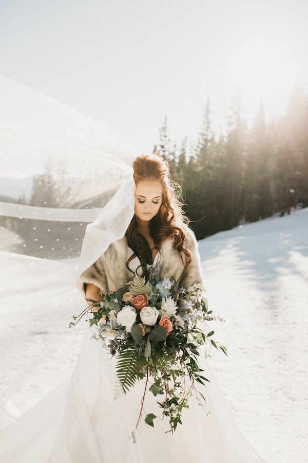 a woman in a wedding dress holding a bouquet and veil over her head while standing in the snow