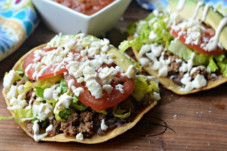 two tacos with lettuce, tomatoes and other toppings next to a bowl of salsa