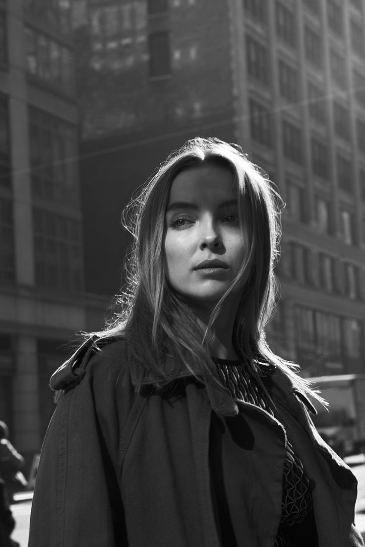 a woman standing in front of a tall building wearing a trench coat and looking at the camera