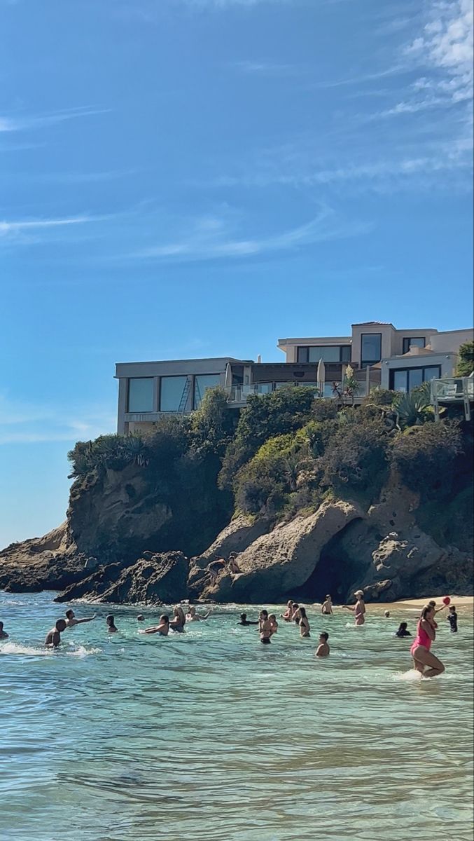 many people are swimming in the ocean near some houses on top of a hill,