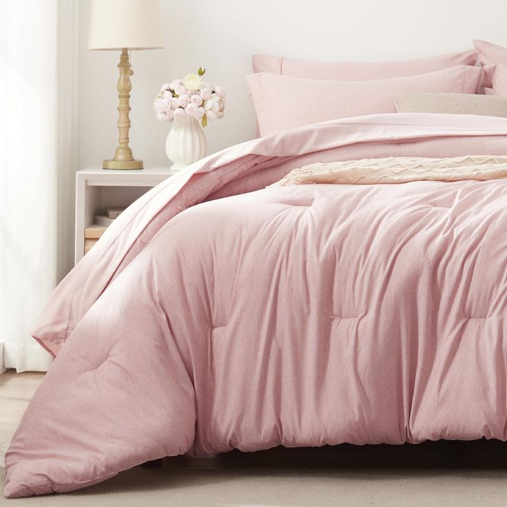 a bed with pink comforter and pillows in a white room next to a lamp