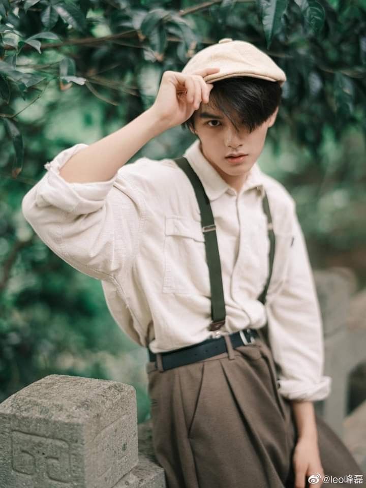 a young man wearing suspenders and a hat standing next to a stone wall with trees in the background