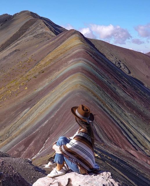 a person sitting on top of a rock next to a rainbow colored mountain range in peru