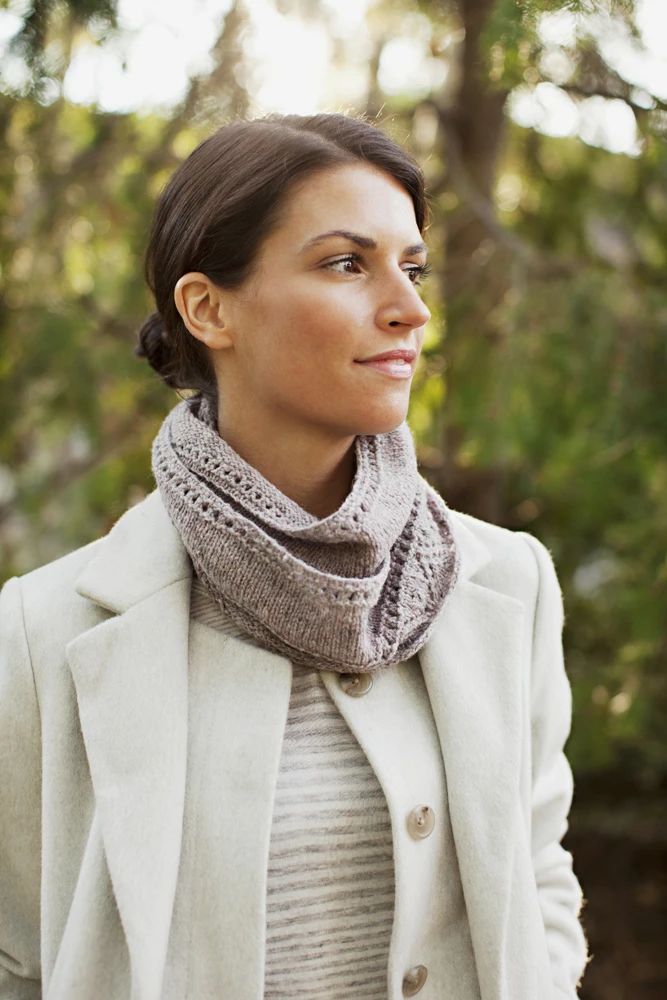 a woman wearing a white coat and gray scarf standing in front of trees with her eyes closed