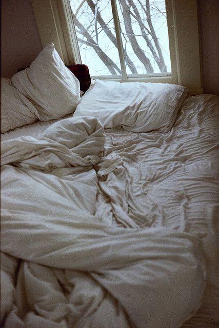 an unmade bed in front of a window with white sheets and pillows on it