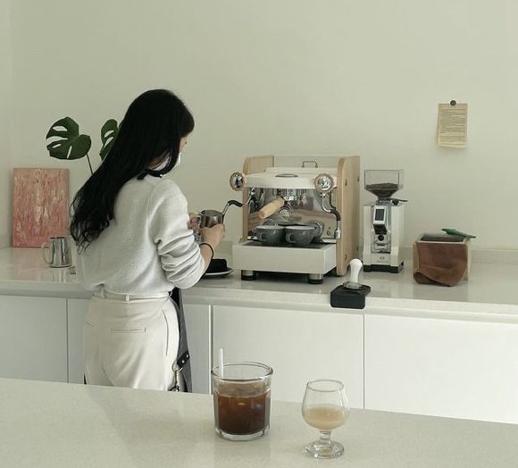 a woman standing in front of a counter next to a glass of liquid and a coffee maker