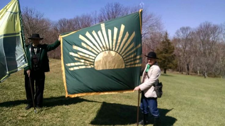 two people holding flags in the grass with trees in the backgrouund and one person standing next to them