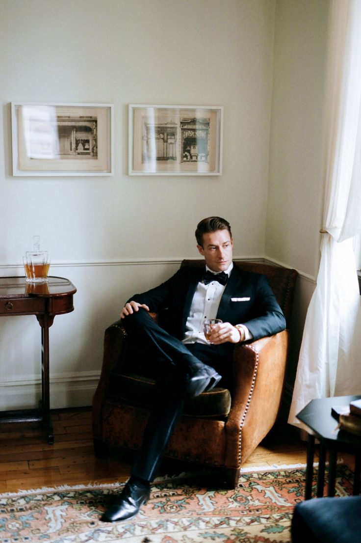 a man in a tuxedo is sitting on a chair and holding a drink