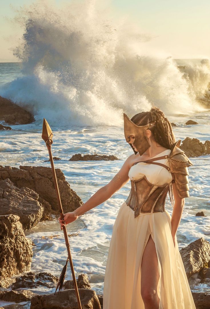 a woman dressed in costume standing on rocks next to the ocean with a spear and shield