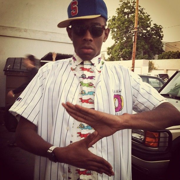 a man wearing a baseball cap and tie standing in front of a car with his hands out