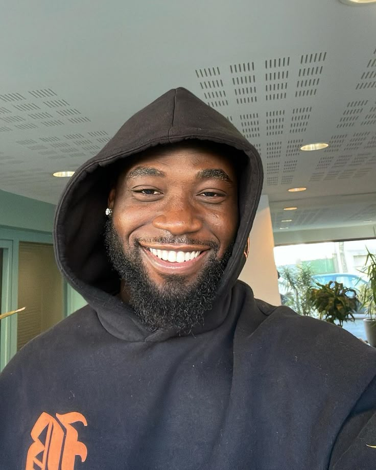 a man with a beard wearing a black hoodie smiles at the camera while standing in an office