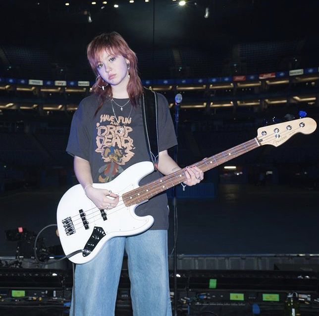 a woman holding a white bass guitar in front of a stage with lights on it