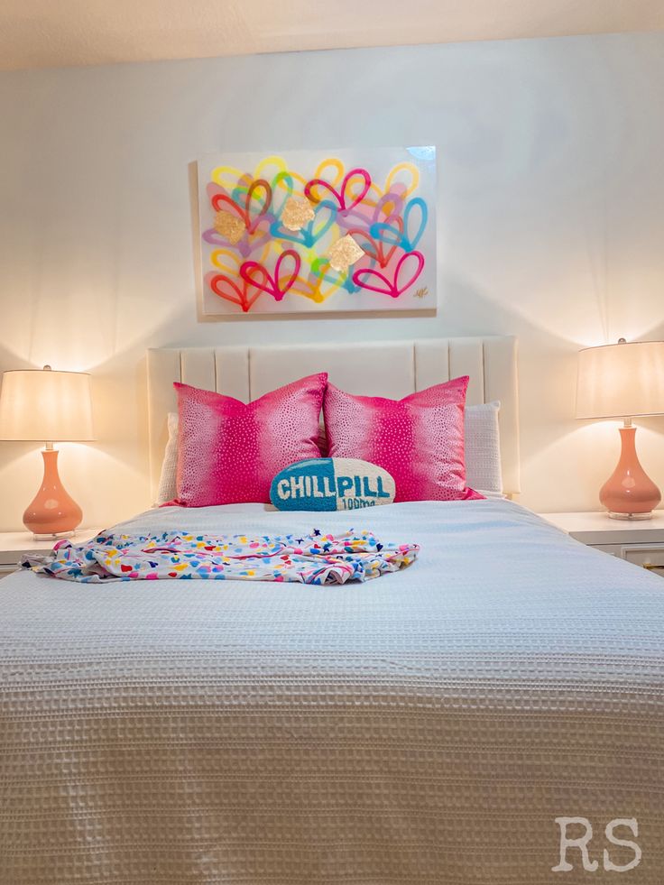 a white bed topped with pillows next to two pink lamps on either side of it