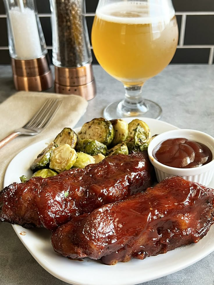 a plate with ribs and brussel sprouts next to a glass of beer