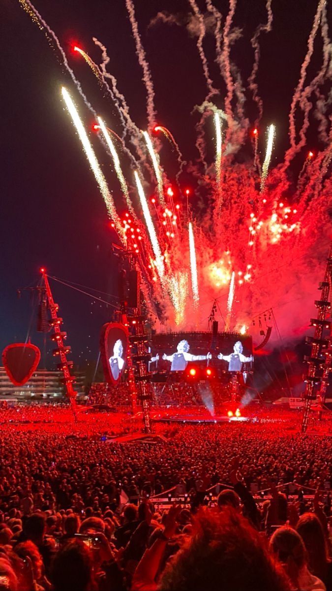 fireworks are lit up in the night sky above an outdoor concert venue with red and white lights